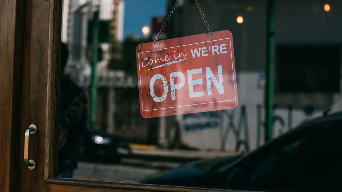 A weathered sign reading Come in We are Open hanging in a shop window at dusk