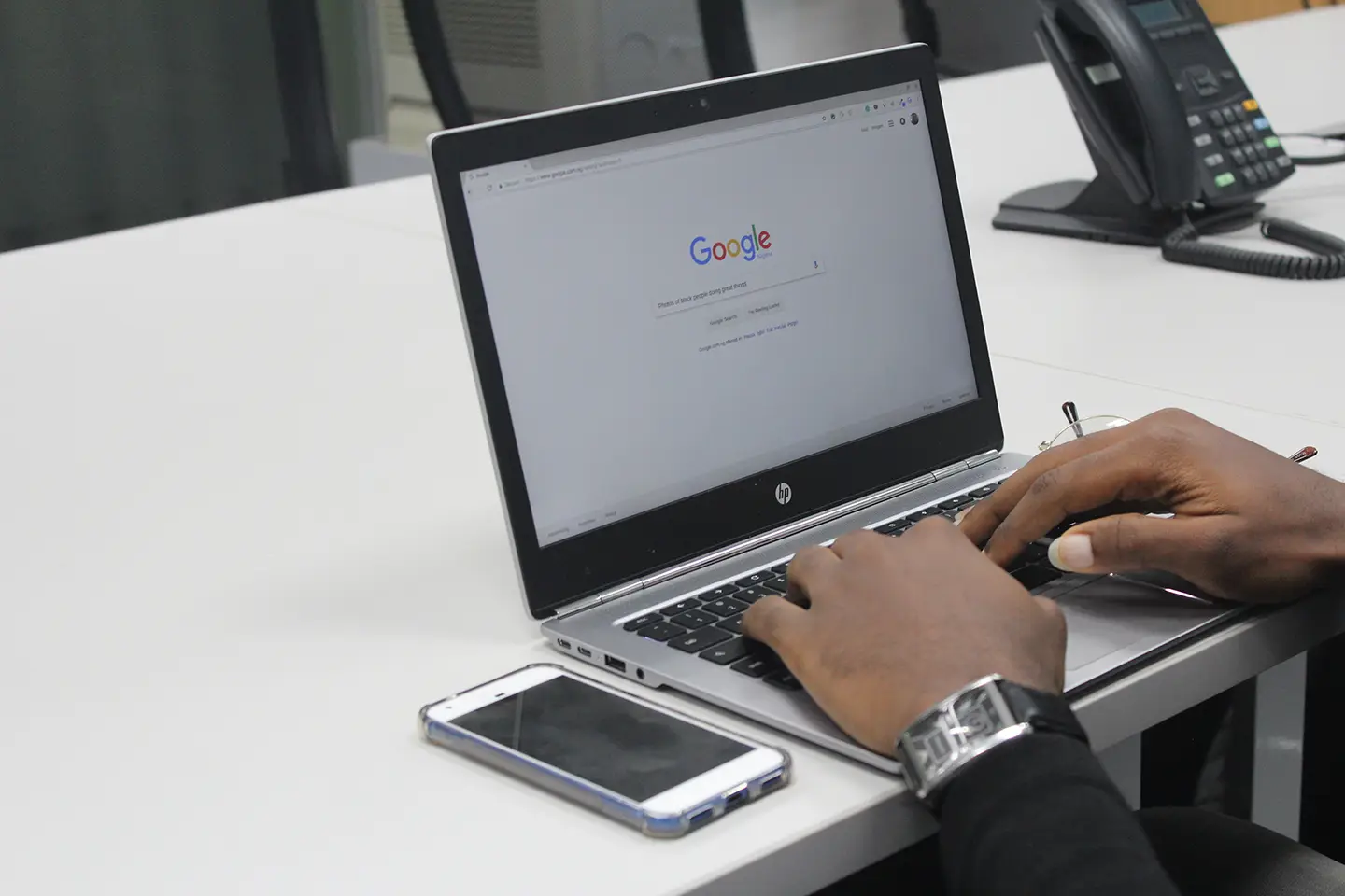 Someone typing on a laptop showing the Google home page, with a phone on the desk beside it