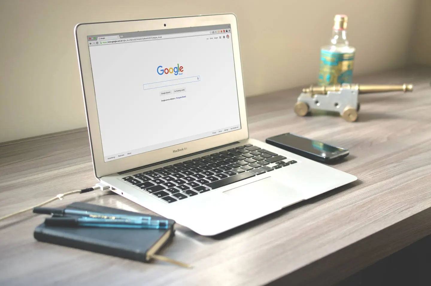A laptop open on a wooden desk showing the Google home page, beside a phone and a notebook