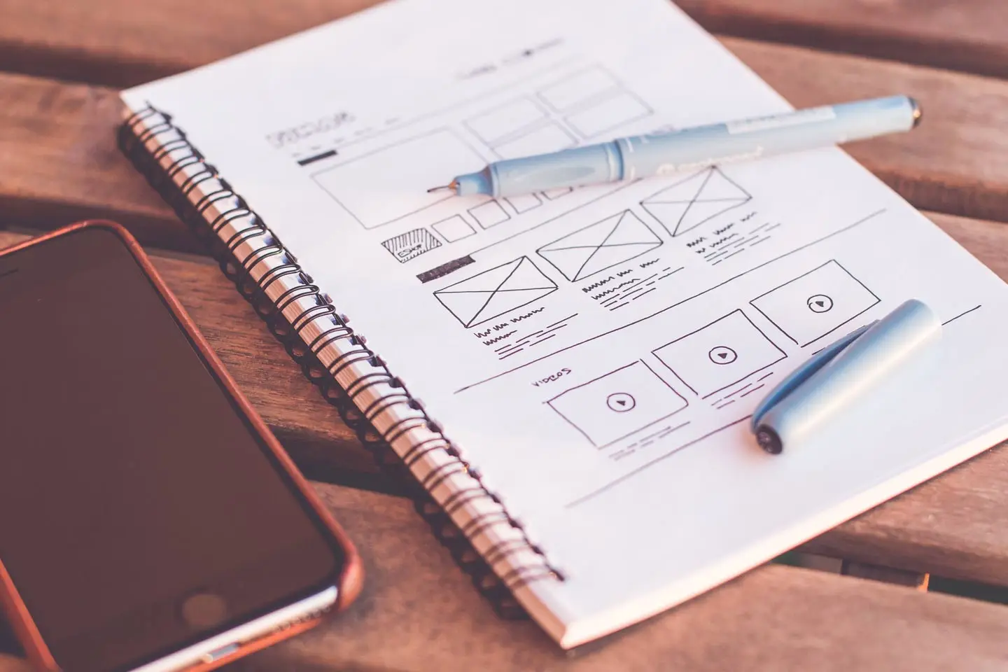 A printed page wireframe on a spiral notebook with two pens and a phone beside it